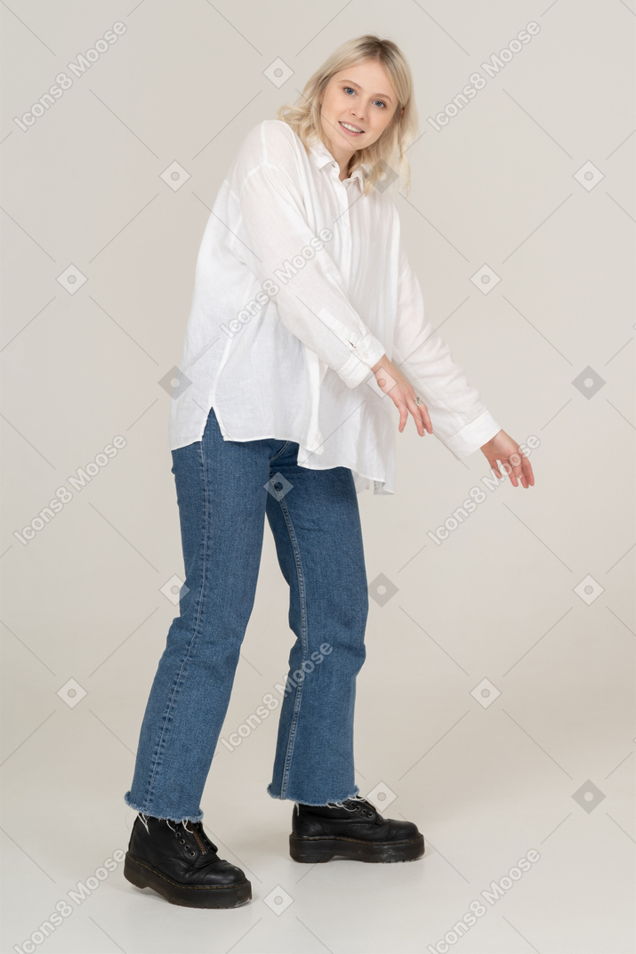 Vista de três quartos de uma mulher loira em roupas casuais, dando um passo para o lado enquanto estende as mãos e olhando para a câmera