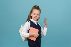 Una colegiala sonriente sosteniendo un libro y saludando