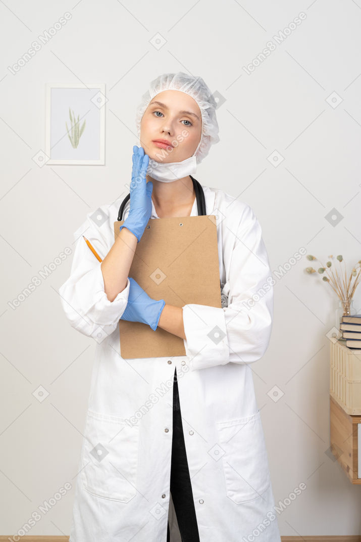 Front view of a pleased young female doctor holding pencil and tablet & touching face
