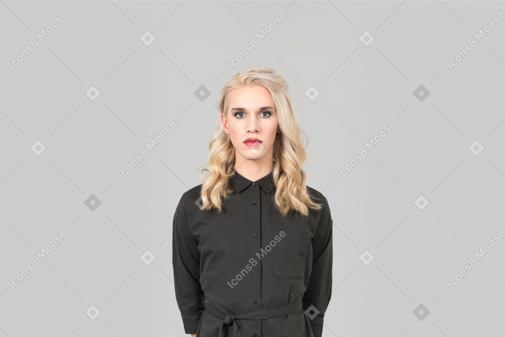 Beautiful trans person in a black dress and with makeup on, standing against the plain grey background, looking forward with a serious face expression