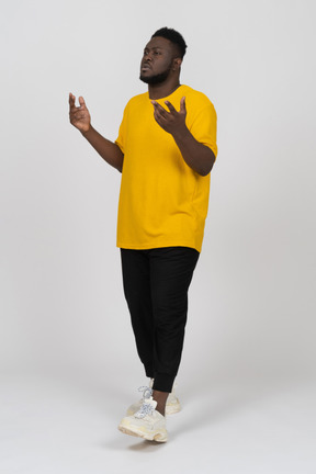 Vista di tre quarti di un giovane uomo dalla pelle scura gesticolante pensieroso in maglietta gialla
