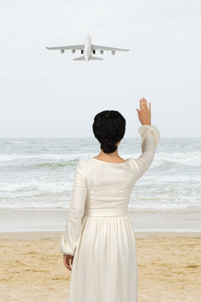 A woman in a white dress standing on a beach and waving to an airplane