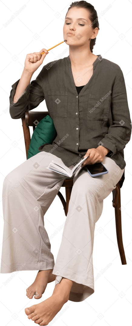 Vista frontal de una mujer joven pensativa vistiendo ropa de casa sentado en una silla con lápiz y cuaderno