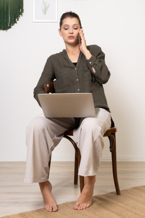 Front view of a busy young woman sitting on a chair with a laptop & mobile