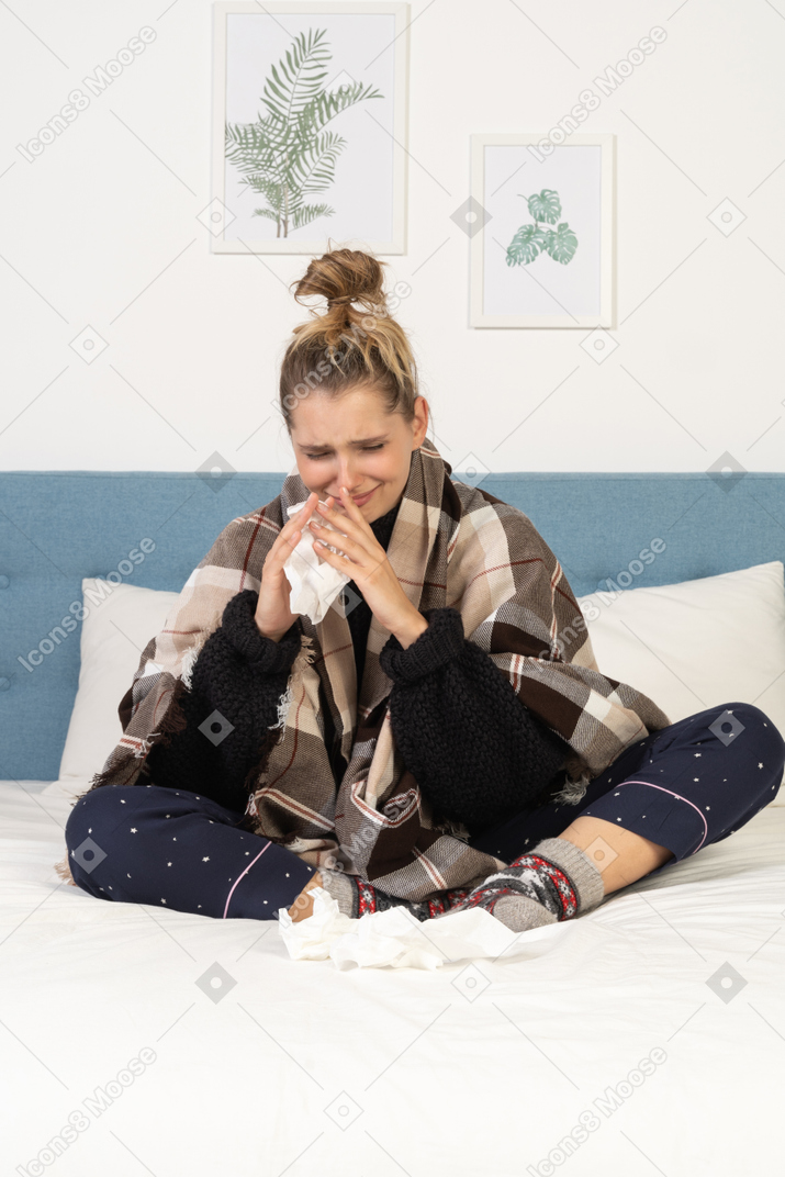 Vista frontal de uma jovem doente de pijama enrolada em um cobertor xadrez na cama assoando o nariz