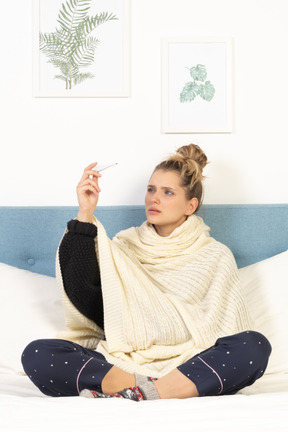 一位裹着白色毯子的困惑的年轻女士坐在床上，拿着温度计的前视图