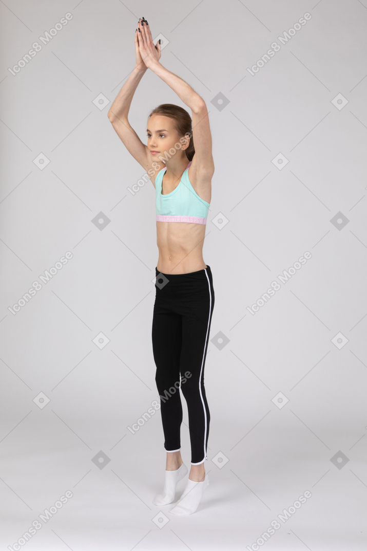 Three-quarter view of a teen girl in sportswear standing on tiptoes and raising hands