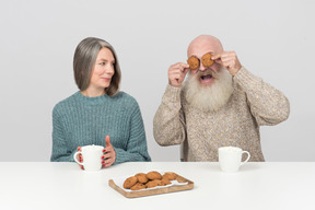 Elder man pretending to have cookies' eyes sitting next to his wife