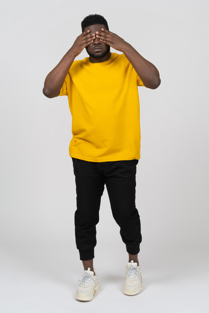 Front view of a young dark-skinned man in yellow t-shirt hiding his eyes