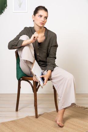 Front view of a thoughtful young woman wearing home clothes sitting on a chair and making notes