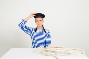 Famale sailor sitting at the table with marine rope on it and holding sailor cap