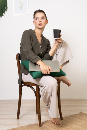 Vista di tre quarti di una giovane donna seduta su una sedia che tiene il suo laptop e tocca una tazza di caffè