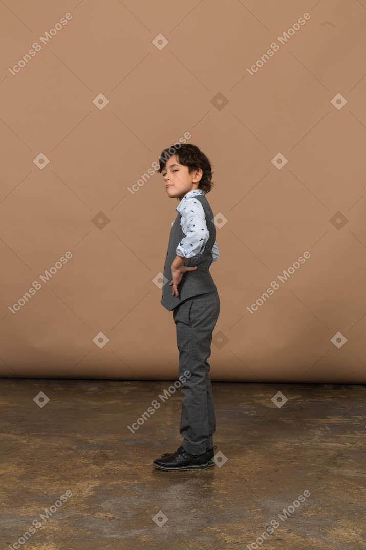 Вид сбоку на симпатичного мальчика в сером костюме, стоящего с руками на бедрах и смотрящего вверх