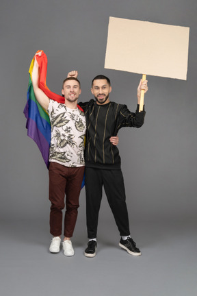 Vista frontal de dos jóvenes sonrientes levantando una valla publicitaria y una bandera lgbt