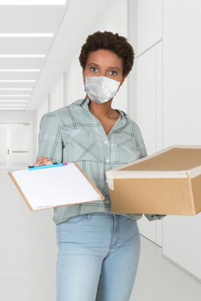 A woman wearing a face mask holding a box and a clipboard