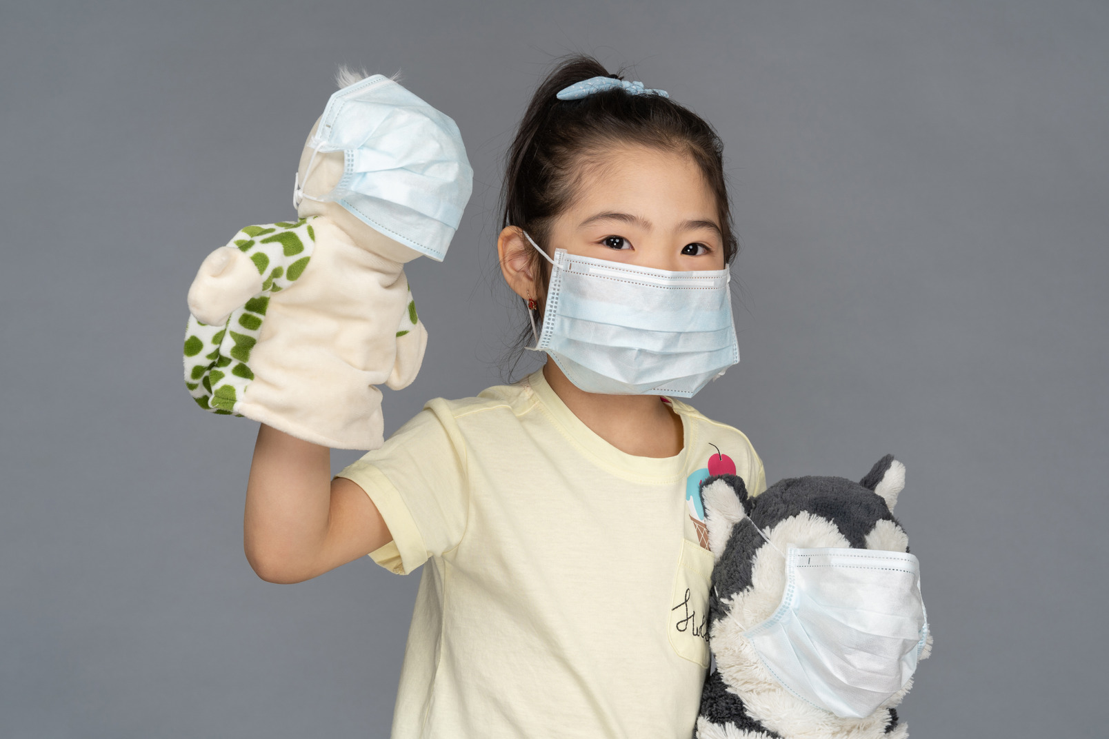 Close-up of a little girl holding two toys, all wearing face masks