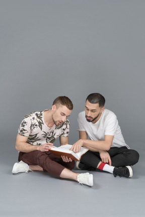 Vorderansicht von zwei jungen männern, die auf dem boden sitzen und ein buch studieren