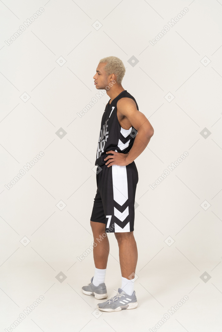 Vista laterale di un giovane giocatore di basket maschile che mette le mani sui fianchi e guarda da parte