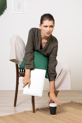 Front view of a confused young woman sitting on a chair and holding her laptop & touching coffee cup