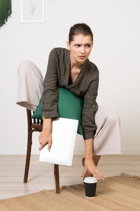 Vue de face d'une jeune femme confuse assise sur une chaise et tenant son ordinateur portable et touchant une tasse de café