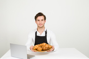 Young baker holding plate of croissants in front of laptop