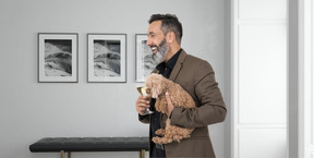 Homme mûr avec une coupe de champagne et un chiot debout devant les affiches dans une galerie, parlant et riant