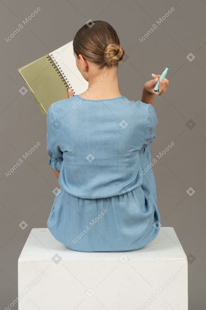 Vista trasera de una joven vestida de azul sentada en un cubo y tomando notas