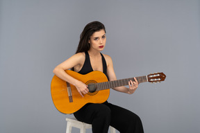 Front view of a sitting young lady in black suit playing guitar