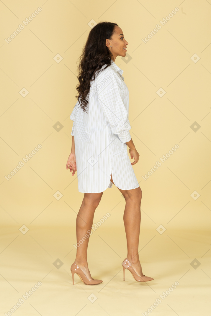 Вид сбоку улыбающейся идущей темнокожей молодой девушки в белом платье