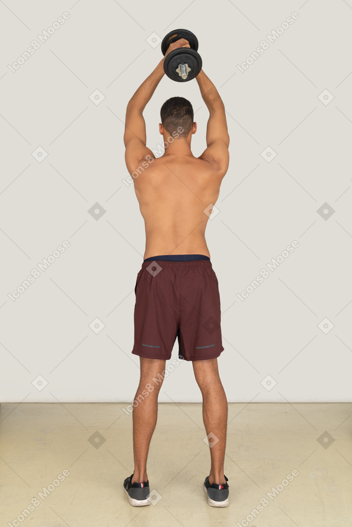 A backside view of the muscular young guy dressed in red shorts and holding the dumbbell above his head