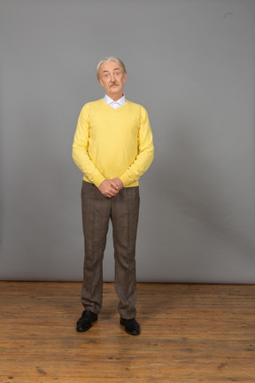 Front view of a confused old man holding hands together and wearing yellow pullover