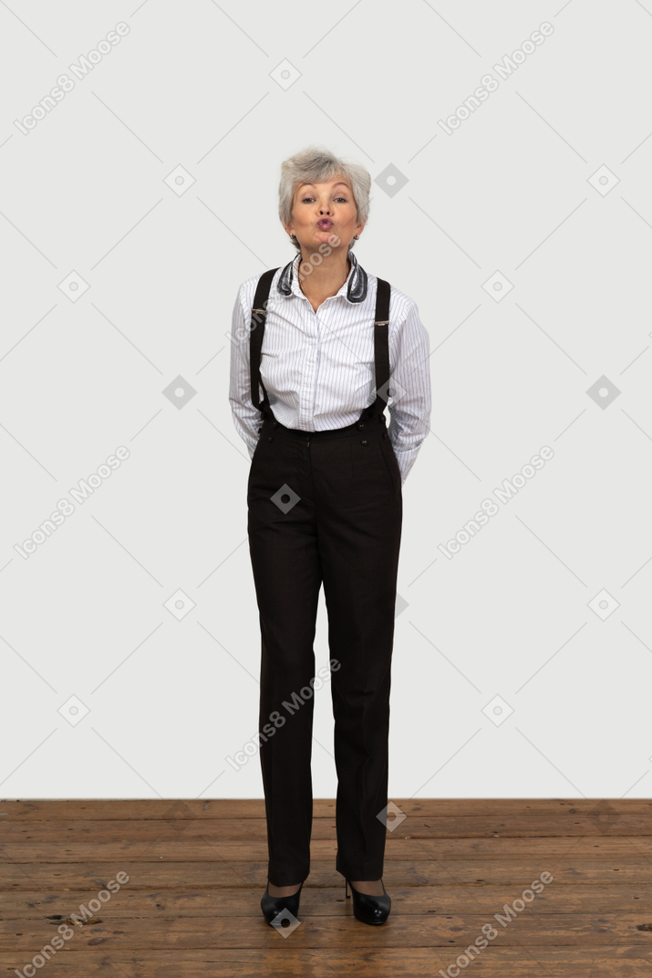 Front view of an old funny female in office clothes grimacing with her hands behind back sending a kiss