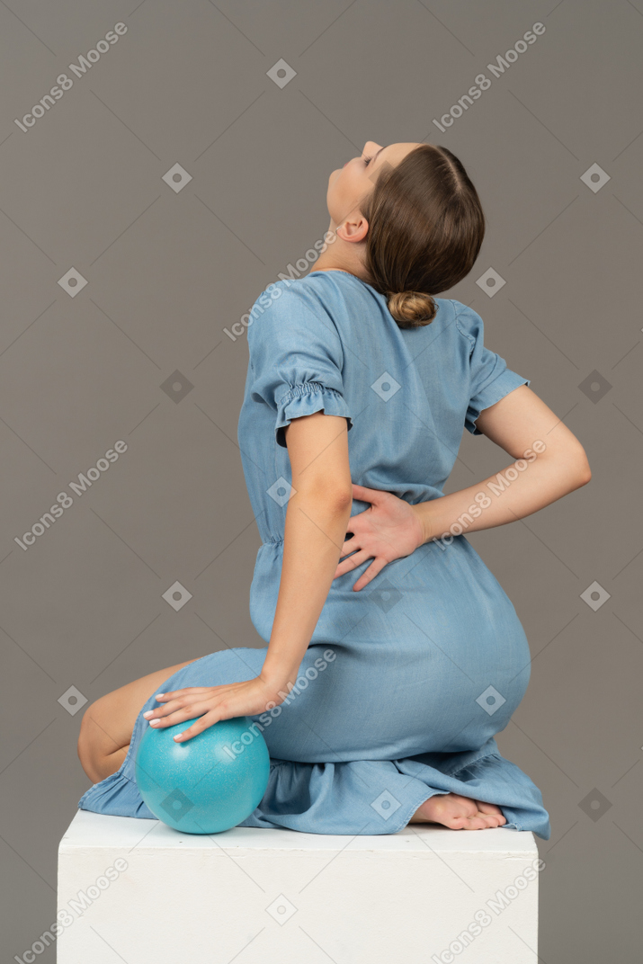 Vue de trois quarts arrière d'une jeune femme assise sur un cube avec une boule bleue