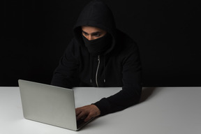 Cara de hacker sentado à mesa e trabalhando no laptop no escuro