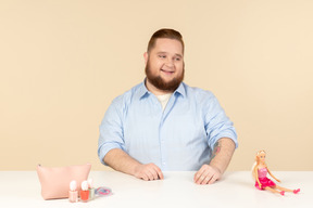 Sonriente tímido joven grande sentado a la mesa y sosteniendo la muñeca barbie