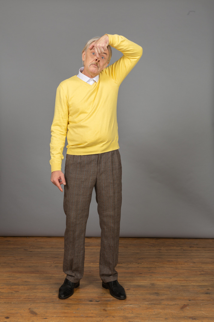Front view of an old funny man in yellow pullover touching nose and looking at camera