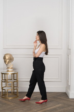 Vista lateral de uma jovem mulher olhando pensativamente de lado enquanto está de pé ao lado da escultura dourada grega