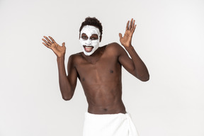 Молодой темнокожий мужчина с белым банным полотенцем на талии занимается уходом за лицом