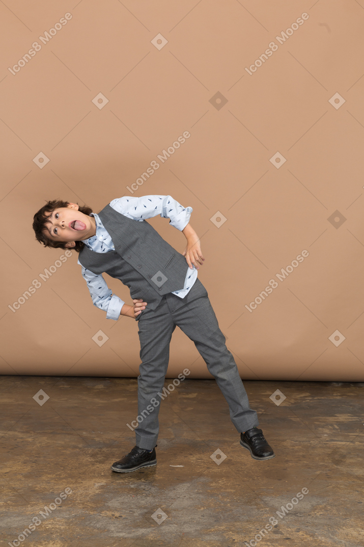 Вид спереди мальчика в костюме, позирующего с руками на бедрах и показывающего язык