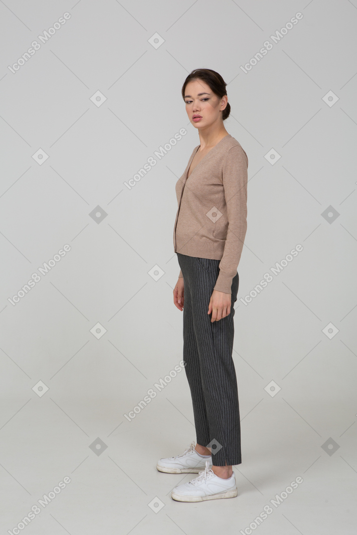 Вид в три четверти угрюмой молодой леди, стоящей неподвижно в пуловере и штанах и смотрящей в сторону