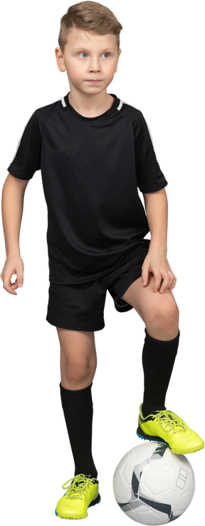 Vista frontal de un niño en uniforme de fútbol poniendo su pie en la pelota y mirando a un lado