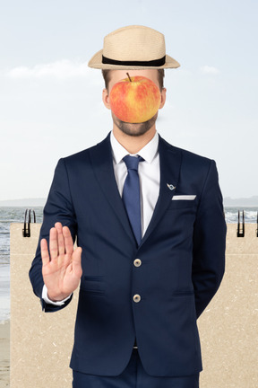A man in a hat with an apple on his face standing in front of a notebook near the sea with palm to the camera