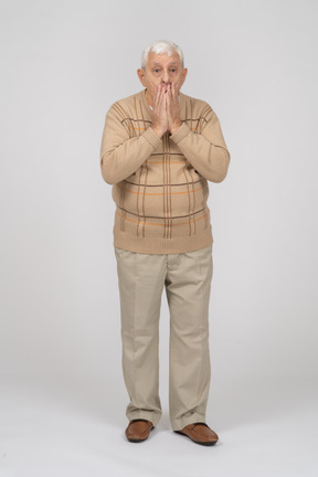 Front view of an impressed old man in casual clothes covering mouth with hands