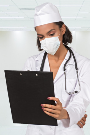 A female doctor in a white lab coat and mask holding a clipboard