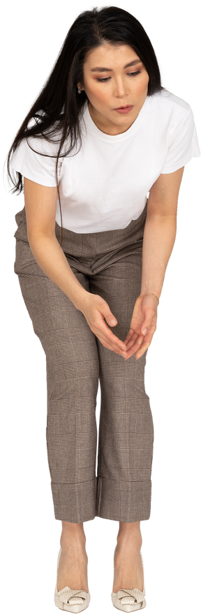 Vista frontale di una giovane donna interrogativa in calzoni e maglietta alzando le mani e chinandosi
