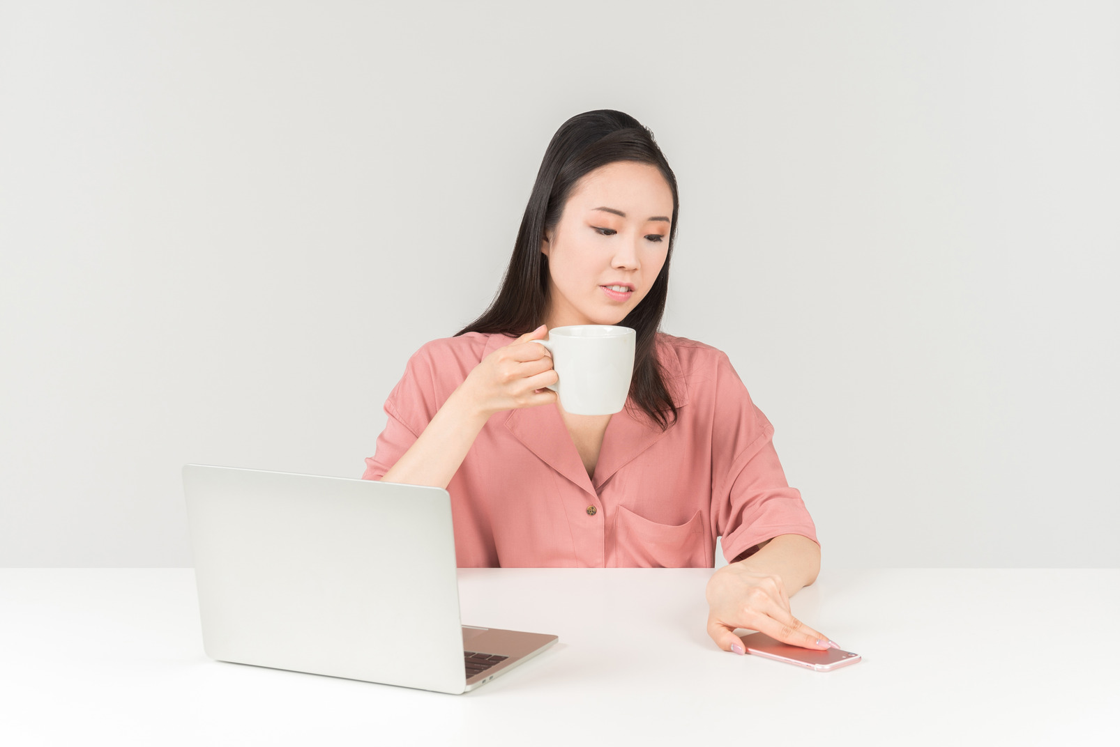 Young asian woman having tea, looking at something and laptop and taking phone