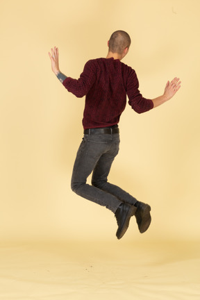 Back view of a jumping young man in red pullover raising his hands