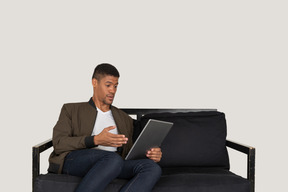 Front view of a shocked young man sitting on a sofa while watching the tablet