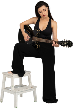一位年轻的女士，穿着黑色西装，抱着吉他，把腿放在凳子上的四分之三的视图