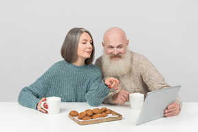 Пожилая пара пьет кофе и смотрит фильм на планшете